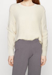 Roomkleurige gebreide trui met een geribbelde textuur en ronde halslijn, gecombineerd met donkergrijze geribbelde broek. Beide hebben een ontspannen pasvorm.