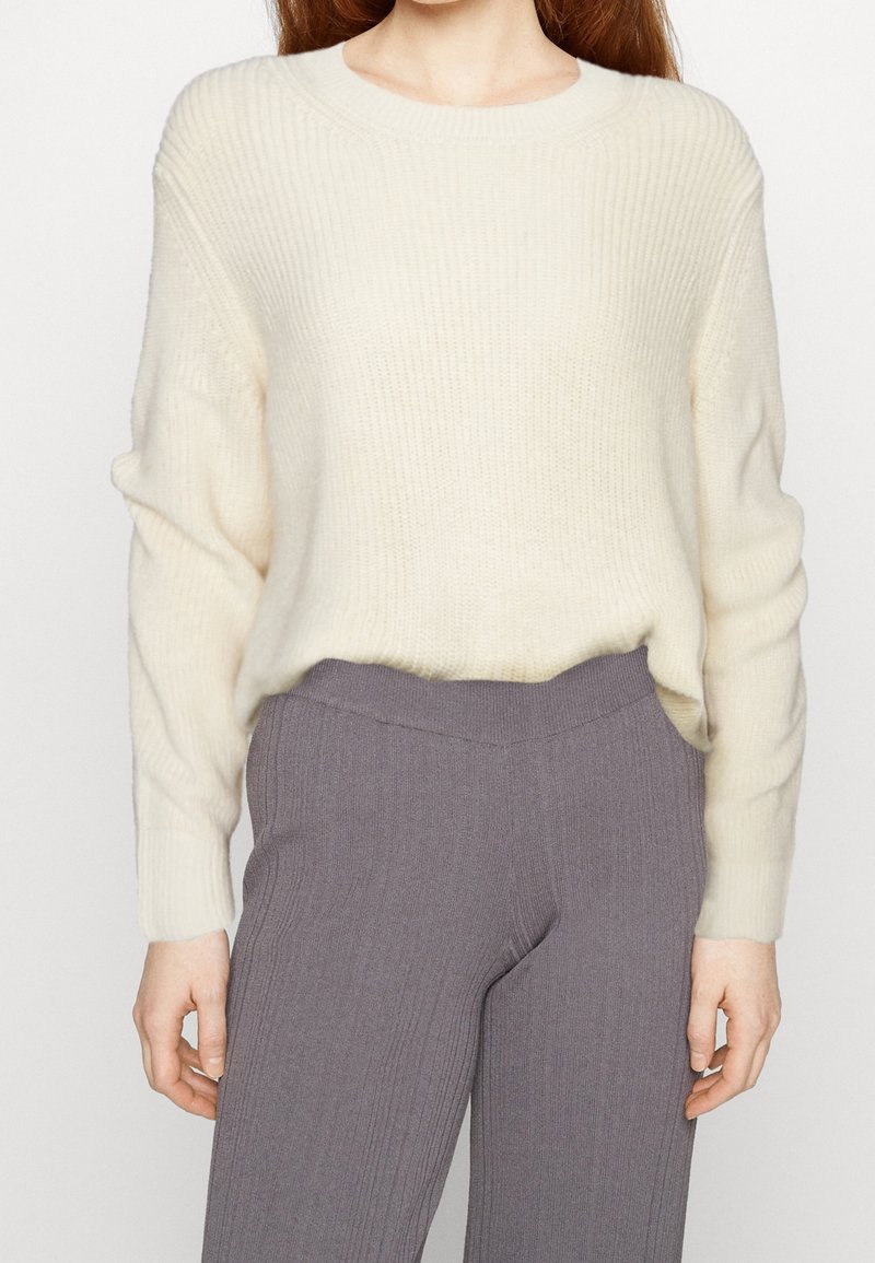 Roomkleurige gebreide trui met een geribbelde textuur en ronde halslijn, gecombineerd met donkergrijze geribbelde broek. Beide hebben een ontspannen pasvorm.