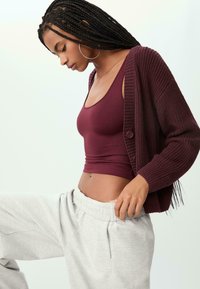 Burgunderrote Strickjacke aus strukturiertem Material und mit Knöpfen, getragen über einem burgunderfarbenen Tanktop, kombiniert mit hellgrauen Sweatpants.
