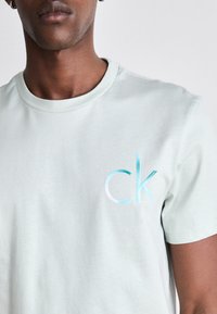 Šviesiai žalia medvilninė marškinėliai su apvaliu kaklu ir subtiliu gradientu mėlynos ir baltos spalvos "ck" logotipu ant krūtinės.