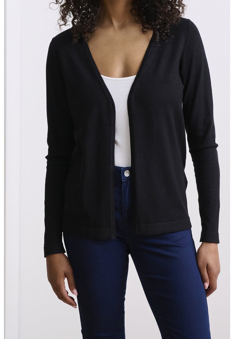 Svart cardigan i lätt tyg, med V-ringning, långa ärmar och öppen framsida. Bärs över en vit topp och kombineras med blå jeans.