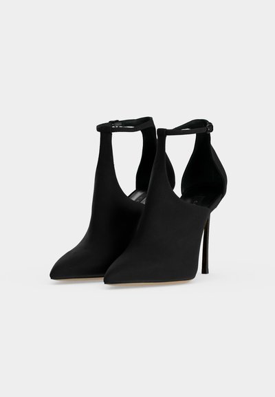 Paire de chaussures à talons noirs à bouts pointus et brides à la cheville, présentées sur un fond gris clair uni.