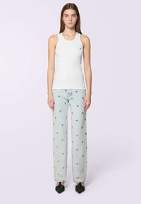 Donna in piedi che indossa un top senza maniche bianco, jeans azzurri chiari con borchie metalliche e scarpe nere con punta. Sullo sfondo, un colore pallido.