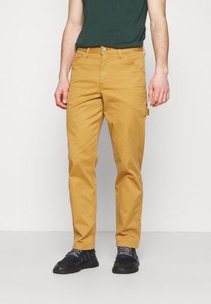 Mann trägt senfgelbe gerade geschnittene Hose, schwarze Sneakers und ein dunkelgrünes Hemd, steht vor einem einfarbigen hellen Hintergrund.