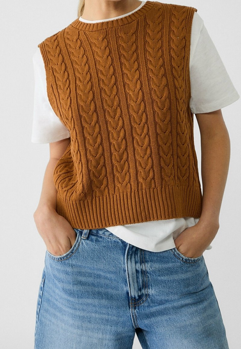 Personne portant un gilet sans manches en tricot torsadé marron sur une chemise blanche à manches courtes et un jean bleu, les mains dans les poches.
