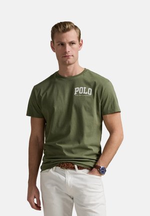 Camiseta de algodón verde oliva con el logo "POLO RALPH LAUREN" en blanco, cuello redondo, mangas cortas, combinada con pantalones blancos y un cinturón marrón.