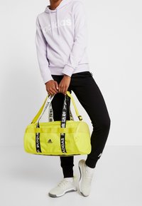 Borsa a busta gialla con accenti neri, dotata di spallacci personalizzati. Il modello indossa un felpa leggera viola e pantaloni neri con scarpe bianche.