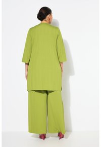 Dlouhý, volný outfit z jasně limetkové látky. Má tříčtvrteční rukávy a široké kalhoty. Oblečené s červenými lodičkami.