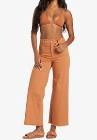 Top de bikini marrón combinado con pantalones naranjas de tiro alto y pernera ancha, con dobladillo deshilachado y textura suave. Lleva sandalias planas marrones.
