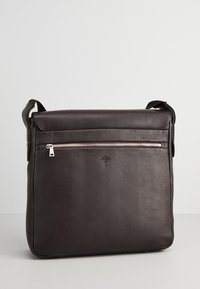 Borsa a spalla in pelle marrone con patta superiore e tasca con zip frontalmente. Presenta una texture liscia e un dettaglio del logo in rilievo sul davanti.