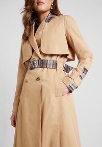 Donna che indossa un trench beige con colletto a quadretti, cintura a quadretti e polsini a quadretti, in piedi con una mano in tasca.