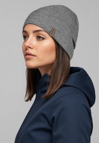 Grijze gebreide muts van zachte stof, met een klein bruin label. Draagt een marineblauwe jas met rits, profielopname van dichtbij.