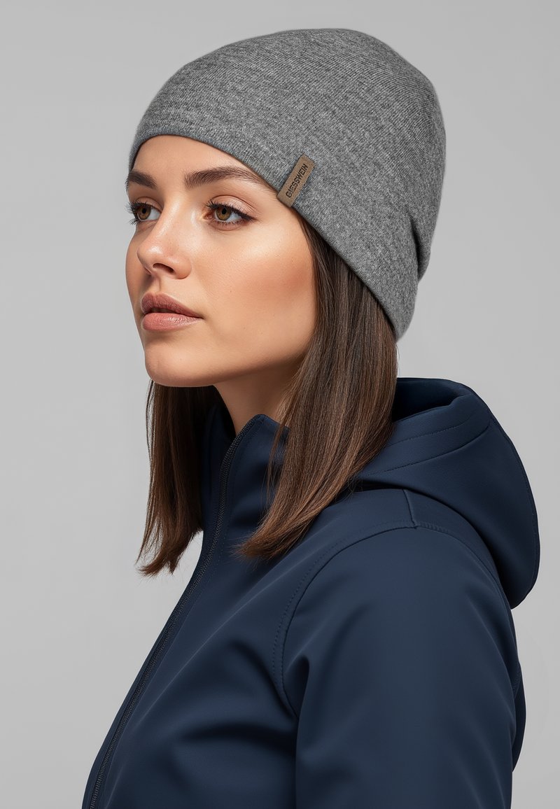 Grijze gebreide muts van zachte stof, met een klein bruin label. Draagt een marineblauwe jas met rits, profielopname van dichtbij.