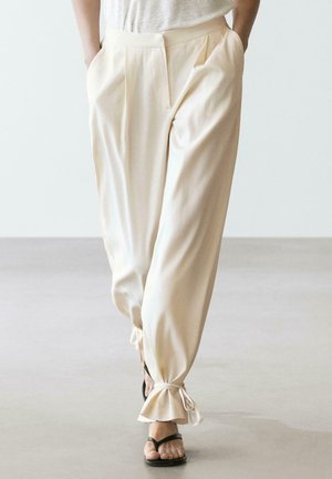 Osoba nosící volné krémové kalhoty se staženými manžetami uvázanými na mašle a černé sandály s otevřenou špičkou, chodící po světlé podlaze.