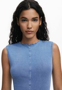 Figurbetonter ärmelloser Oberteil aus hellblauem Rippstoff mit einem gewellten Ausschnitt und sichtbaren Mittelnahtdetails. Glatte Textur über das ganze Stück.