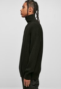 Pull col roulé noir, texturé, à coupe décontractée et détails côtelés, avec des manches longues et un col haut, vu de côté.