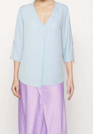 Lichtblauwe v-halsblouse met driekwartmouwen, gestructureerde stof en een overlappend ontwerp aan de voorkant. Gepaarde met een vloeiende lavendelkleurige rok.