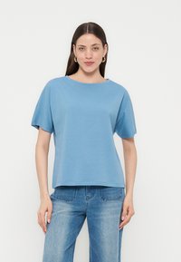 Γυναίκα με μακριά καστανά μαλλιά που φορά χρυσούς κρίκους σκουλαρίκια, ένα γαλάζιο κοντομάνικο πουκάμισο και μπλε τζιν, στέκεται μπροστά σε λευκό φόντο.