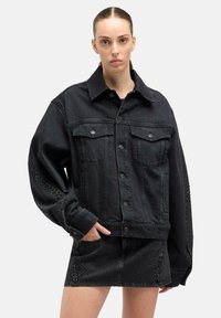 Kobieta z zaczesanymi do tyłu włosami, ubrana w oversize'ową czarną kurtkę z denimu i do tego dopasowaną, ozdobioną ćwiekami mini spódnicę, pozuje z jedną ręką w kieszeni.