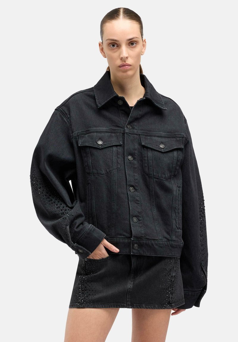 Kobieta z zaczesanymi do tyłu włosami, ubrana w oversize'ową czarną kurtkę z denimu i do tego dopasowaną, ozdobioną ćwiekami mini spódnicę, pozuje z jedną ręką w kieszeni.