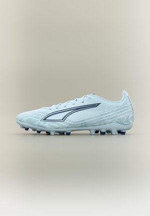 Crampon de football bleu clair avec logo bleu foncé, design à lacets et crampons moulés sur la semelle, présenté de profil sur un fond beige.