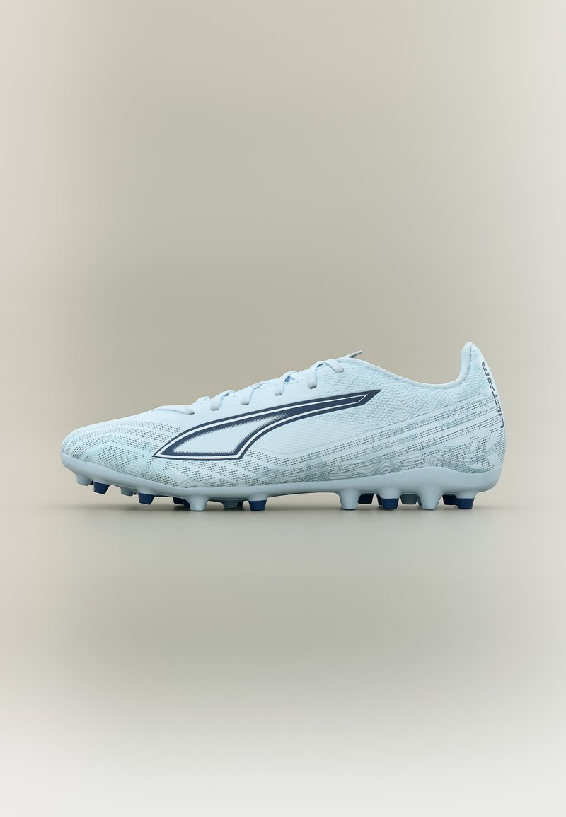 Svetlo modri nogometni čevelj s temno modrim logotipom, z zavezovanjem na vezalke in oblikovanimi kramponi na podplatu, prikazan s strani na bež ozadju.