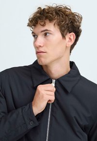 Young man with curly hair zipping up a black collared jacket, looking to his left against a plain light background.
