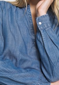 Camicia in denim chiaro con scollo a V, chiusura a bottoni ai polsini e maniche lunghe plissettate. Il tessuto ha una texture liscia e una tonalità di blu sbiadito.