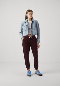 Jeans-gekürzte Jacke, blau; weißes enges Oberteil; bordeauxfarbene Jogginghose mit Kordelzug; hellblaue Sneaker mit dunklen Akzenten.