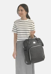 Zaino trapuntato nero con due manici superiori e tasche laterali. Texture liscia e dettagli in cerniera argentata. Indossato da una persona con una camicia a righe e una gonna.