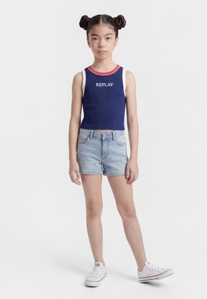 Noor tüdruk kahe kukliga soengu ja tumesinise varrukateta toppi, millel on tekst "REPLAY", hele teksapükslühike ja valged spordijalatsid, seistes.