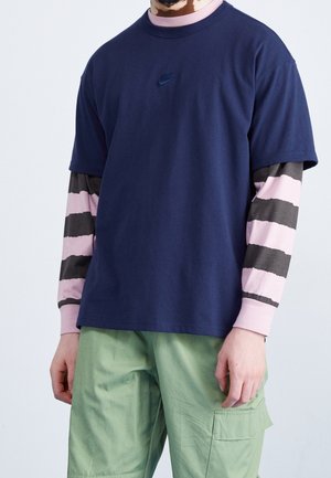 Mężczyzna w granatowej koszulce Nike na różowo-czarnym paskowanym longsleeve'u oraz jasnooliwkowych spodniach typu cargo, stojący na jednolitym tle.