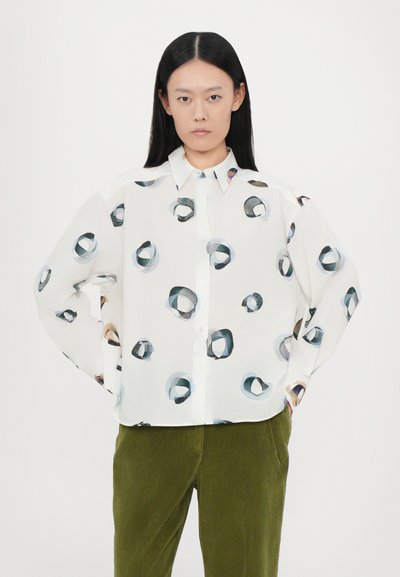 Camisa blanca de botón con un patrón abstracto circular en azul y beige. Presenta un cuello estándar y un corte holgado. Combinada con pantalones de pana verde.