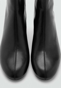 Botas de cuero negras hasta el tobillo con una textura suave, puntera redondeada y detalles de costura lateral. El diseño es elegante y ajustado al pie.