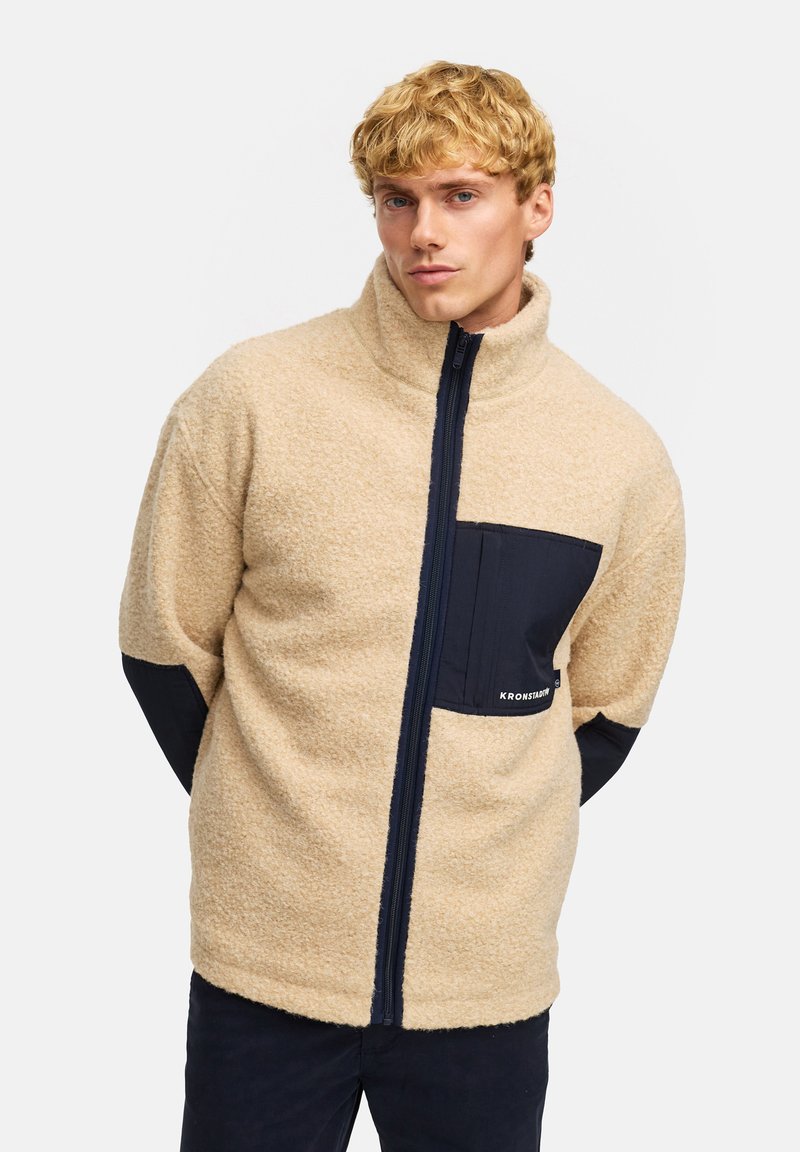 Beige fleecejacka med hög krage, svarta accenter vid axlarna och bröstfickan samt framdragkedja i mitten. Texturerad tygfinish.