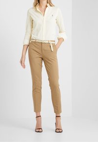 Světle žlutá košile na knoflíky, béžové kalhoty na míru s pleteným bílým pásem a hnědé sandály na podpatku, vystavené na světlém pozadí.