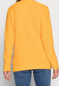 Kirkas keltainen ribattu neuletakki, jossa on pitkät hihat ja sivurei'itykset, sekä pienet reijät hihoissa ja rento istuvuus.