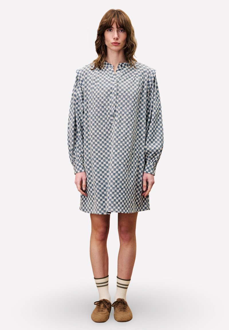 Hemdblusenkleid mit einem blau-weißen Karomuster, Ballonärmeln und einer Länge bis zur Mitte des Oberschenkels. Getragen mit beigen Schuhen und gestreiften Socken.