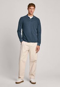 Långärmad marinblå pikétröja i mjukt bomullstyg, kombinerad med ljusbeige byxor och vita sneakers. Enkel design med krage.