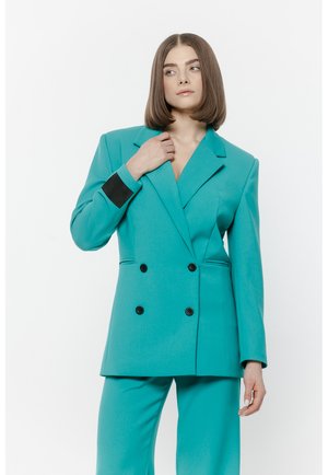 Kobieta o prostych brązowych włosach ubrana w turkusową dwurzędową marynarkę i pasujące spodnie, pozująca na białym tle.