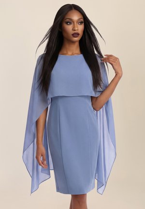 Frau mit langen schwarzen Haaren, die ein hellblaues knielanges Kleid mit fließenden, kapuzenartigen Ärmeln trägt, posiert vor einem schlichten beigen Hintergrund.