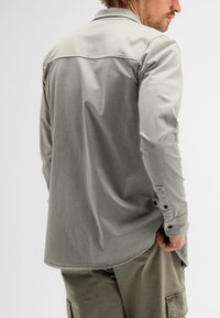 Hellgraues Langarmhemd mit Kragen, Knopfmanschetten und einem leicht abgerundeten Saum. Getragen mit olivgrünen Cargo-Shorts.