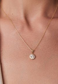 Złoty naszyjnik z łańcuszka z wisiorkiem w kształcie kwiatu, w którym znajduje się centralny przezroczysty kamień otoczony mniejszymi kamieniami, ukazujący misterną dekorację.