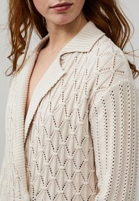 Gilet en tricot de couleur beige clair avec un motif diamant texturé, un col châle côtelé et une conception ouverte.