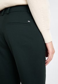 Ciemnozielone dopasowane spodnie z tylnią kieszenią i małym logo. Gładka tekstura, dopasowany krój oraz wysokiej jakości materiał.