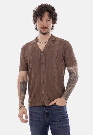 Homme aux cheveux bouclés portant une chemise marron à manches courtes boutonnée et un jean bleu, exhibant des tatouages sur les deux avant-bras, debout avec une main dans la poche.