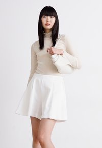 Bílá krátká sukně spárovaná s průhledným béžovým topem s dlouhým rukávem. Modelka drží bílou kabelku se strukturovaným designem. Jednoduchá, moderní estetika.