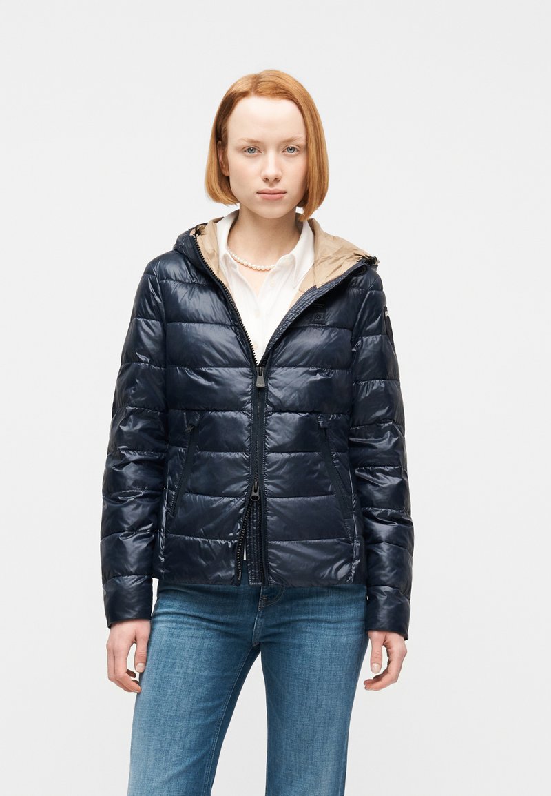 Γυναίκα με κοντά κόκκινα μαλλιά, ντυμένη με μπουφάν navy blue με καπιτονέ υφή, λευκό πουκάμισο με γιακά, κολιέ μαργαριτάρι και μπλε τζιν, σε απλό φόντο.