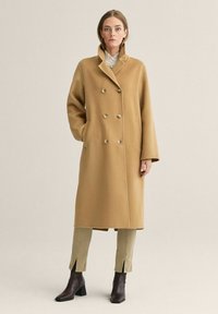 Dobbeltbrystet kasketfrakke i kamelfarve med store knapper, bred krave, sidelommer og en skræddersyet pasform. Båret over beige bukser og mørke hælede støvler.