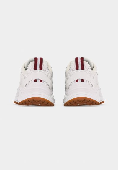 Paire de baskets blanches avec des bandes verticales rouges sur les languettes des talons et des semelles couleur gomme, vues de dos sur un fond clair uni.